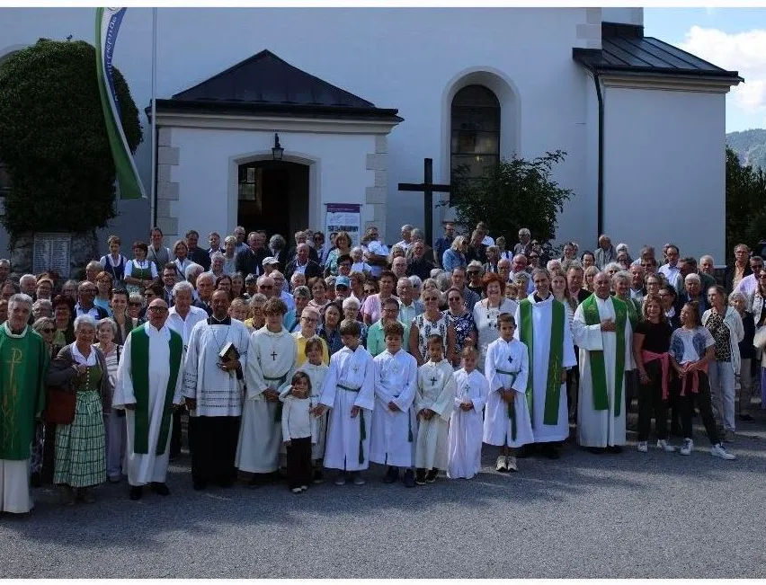 Eine große Gruppe von Menschen, einschließlich Geistlicher und Kinder, versammelt sich vor einer Kirche. Sie sind in weißen und grünen Gewändern gekleidet und einige halten Bücher. Die Kirche hat ein Kreuz auf der Spitze und eine Flagge auf der linken Seite.