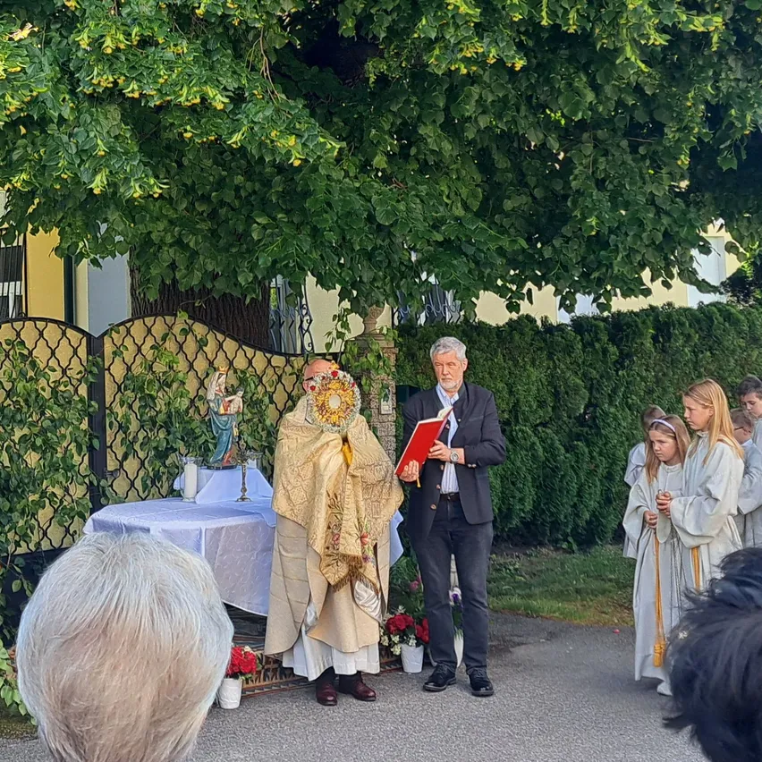 Ein Priester steht an einem Tisch mit Blumen und einer Statue. Ein Mann liest aus einem Buch. Zwei Mädchen in weißen Gewändern stehen hinter ihm. Menschen beobachten unter einem Baum.