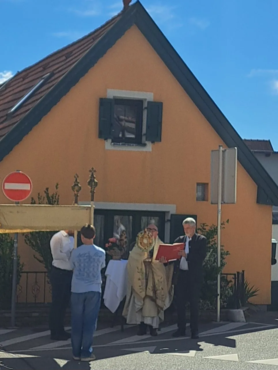 Eine religiöse Zeremonie findet vor einem Gebäude mit Fenster und einem Kreuz auf der Spitze statt. Ein Mann hält ein rotes Buch, während andere in der Nähe stehen.