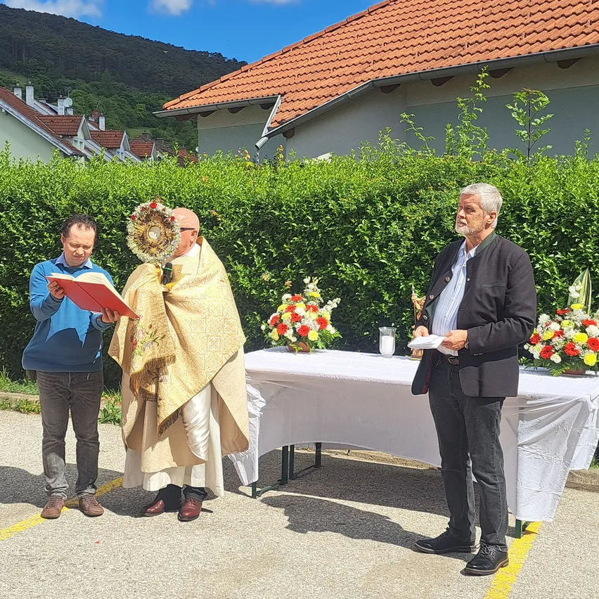 Ein Priester hält ein Buch, während ein anderer aus einem Papier liest. Ein dritter Mann steht neben einem Tisch mit Blumen.