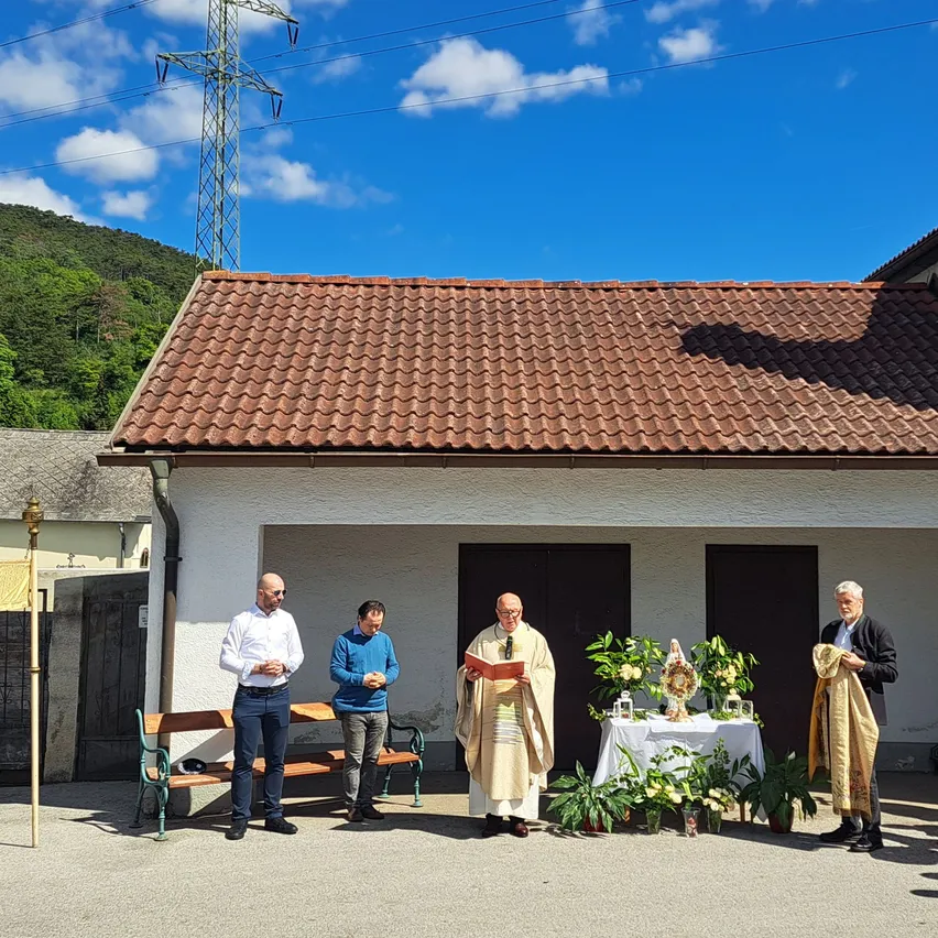 Eine Gruppe von Männern steht vor einer Kapelle, ein Priester liest aus einem Buch. Eine Bank und Blumen sind im Vordergrund.