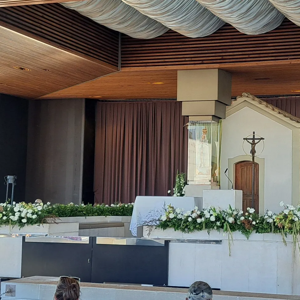 Eine Kapelle mit einem Kruzifix und Blumen davor, mit einer hölzernen Decke und Vorhängen.