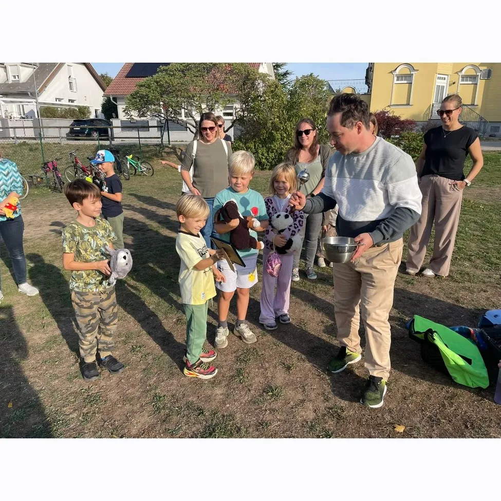 Eine Gruppe von Kindern und Erwachsenen steht auf einem Feld. Der Mann vorne hält ein Kuscheltier und scheint mit den Kindern zu sprechen.