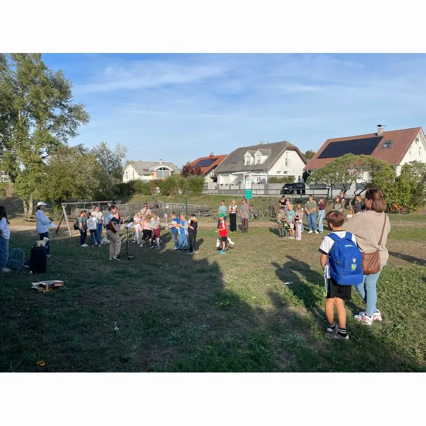 Eine Gruppe von Menschen versammelt sich auf einem Grasfeld, einige stehen und einige sitzen. Im Hintergrund befinden sich Häuser und ein klarer blauer Himmel.
