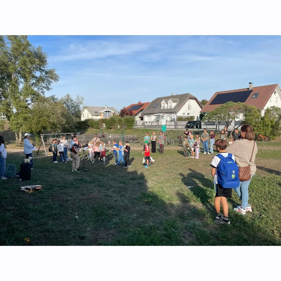 Eine Gruppe von Menschen versammelt sich auf einem Grasfeld, einige stehen und einige sitzen. Im Hintergrund befinden sich Häuser und ein klarer blauer Himmel.