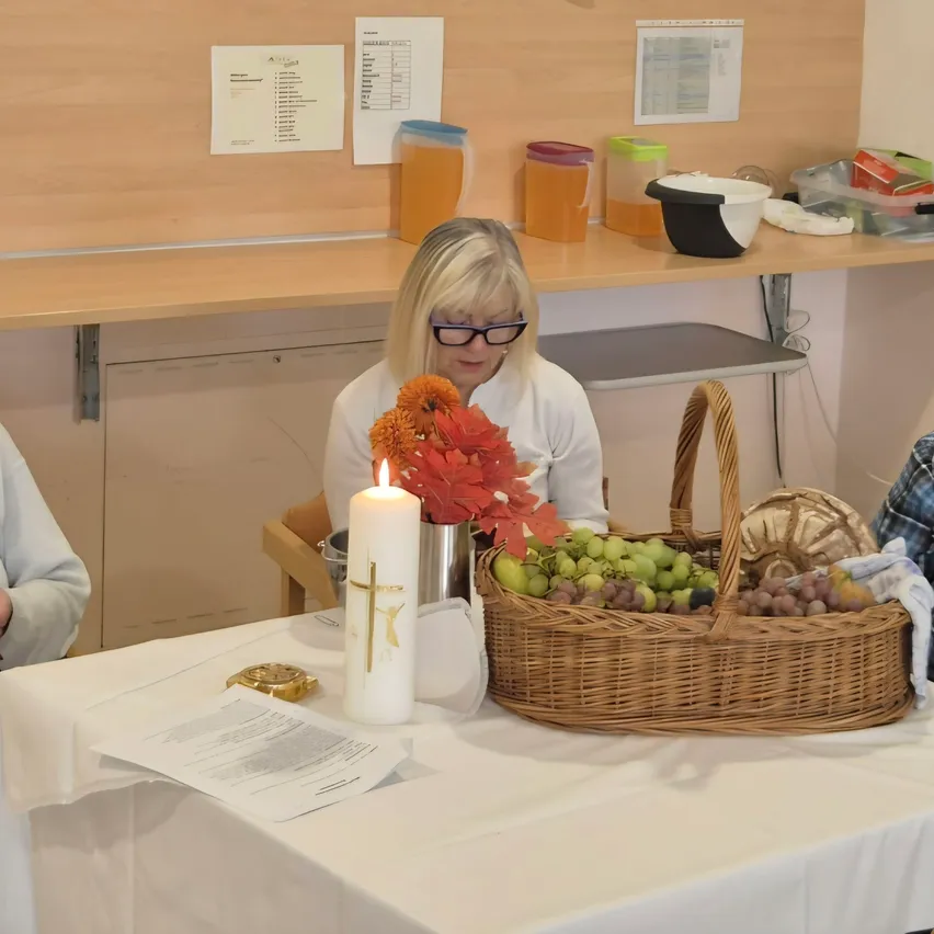 Vier Frauen sitzen um einen Tisch in einem Raum, möglicherweise für eine Besprechung. Der Tisch hat eine weiße Tischdecke, eine Kerze, einen Korb mit Früchten und ein Buch. Die Frau auf der linken Seite liest ein Buch, während die Frau in der Mitte die Frau auf der rechten Seite ansieht.