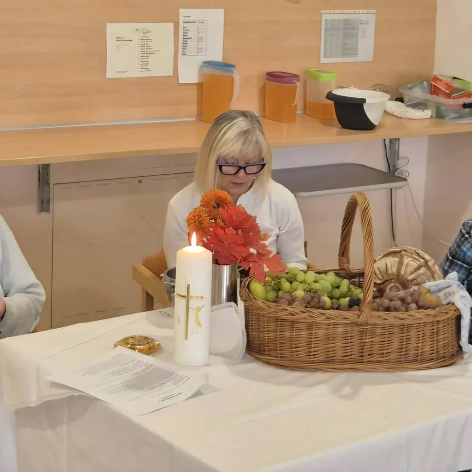 Vier Frauen sitzen um einen Tisch in einem Raum, möglicherweise für eine Besprechung. Der Tisch hat eine weiße Tischdecke, eine Kerze, einen Korb mit Früchten und ein Buch. Die Frau auf der linken Seite liest ein Buch, während die Frau in der Mitte die Frau auf der rechten Seite ansieht.