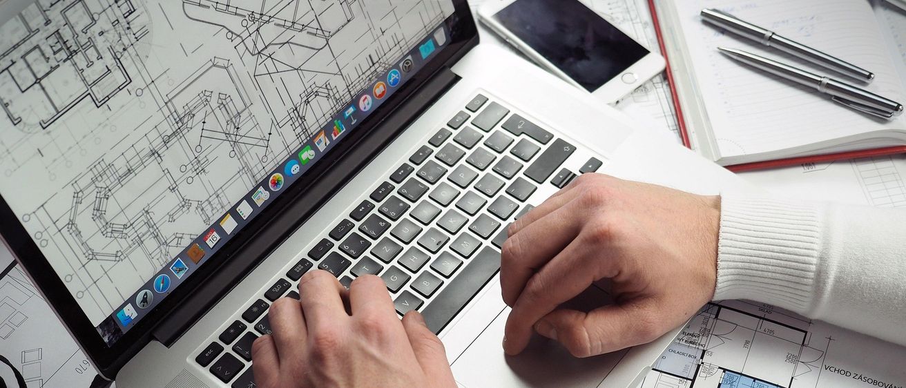 Eine Person, die einen weißen Pullover und eine Uhr trägt, tippt auf einem Laptop mit Bauplänen auf dem Schreibtisch. Ein Handy und ein Notizbuch mit Stiften sind ebenfalls auf dem Schreibtisch.