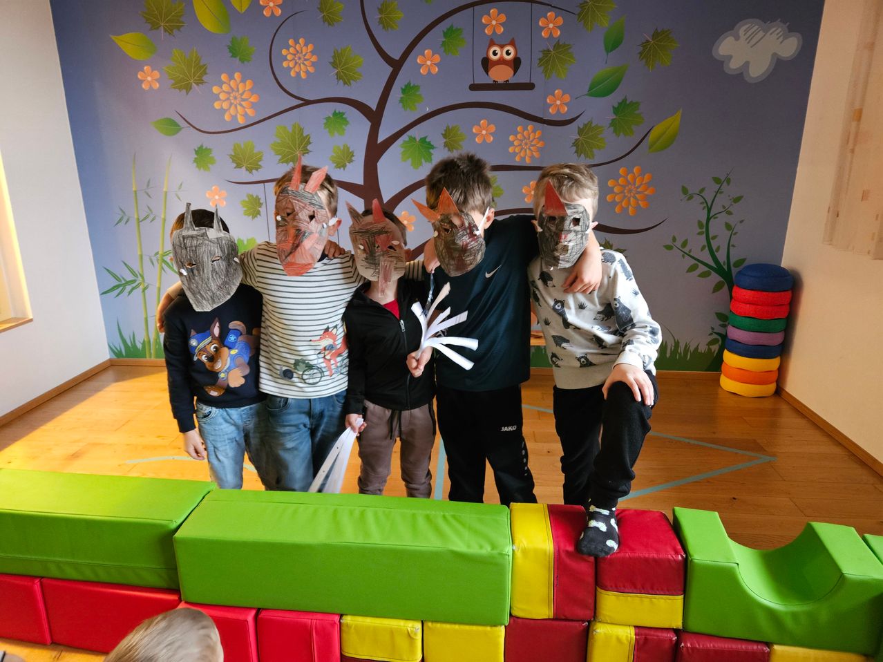 Fünf Kinder stehen zusammen und tragen Tiermasken. Sie befinden sich in einem Raum mit einem farbenfrohen Boden aus gestapelten Blöcken. Hinter ihnen ist eine Wandmalerei mit einem Baum, Blumen und einer Eule.