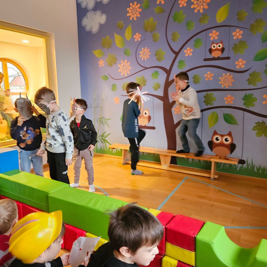 Eine Gruppe von Kindern spielt in einem Raum mit einem Wandbild eines Baumes mit Eulen und Blumen. Einige tragen Masken und Helme, andere klettern auf eine Bank. Ein Junge liest ein Papier.