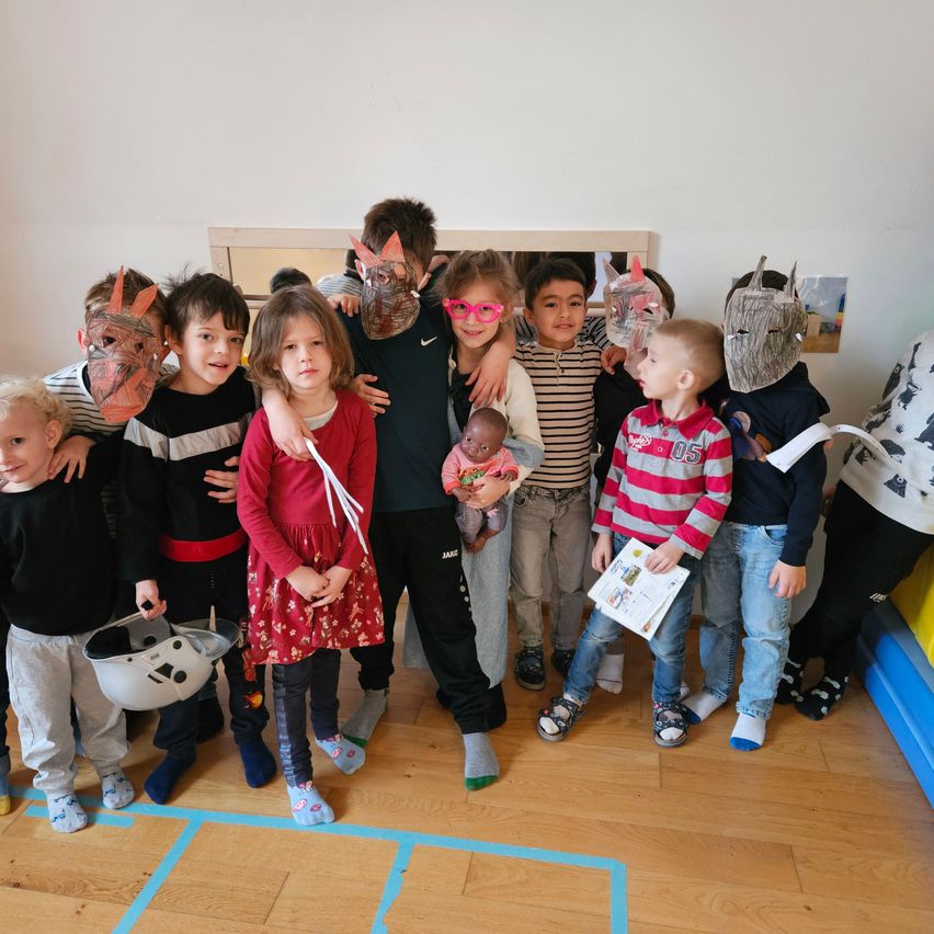 Eine Gruppe von Kindern mit Tiermasken und einem Baby in der Hand steht in einem Raum, einige halten Requisiten.