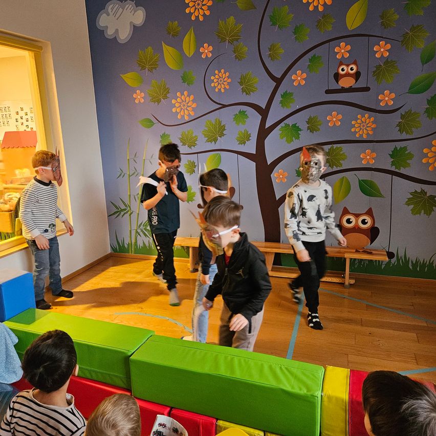 Mehrere Kinder tragen Masken und spielen ein Spiel in einem Raum mit Holzboden. Einige stehen auf einer bunten Matte. Die Wand hinter ihnen ist mit Blättern, Blumen und einer Eule verziert.
