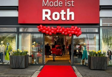 Der Eingang eines Geschäfts namens 'Mode ist Roth' mit rotem Teppich und roten Luftballons. Im Inneren gehen Menschen auf einem Holzboden, und Mannequins zeigen Kleidung. Das Geschäft hat Glaswände und Türen.