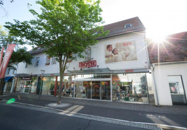 Ein weißes Gebäude mit einem Baum davor zeigt ein Poster eines Paares. Der Text 'ROTH' ist über dem Eingang prominent angebracht. Die Sonne scheint hell und wirft Schatten auf die Straße.