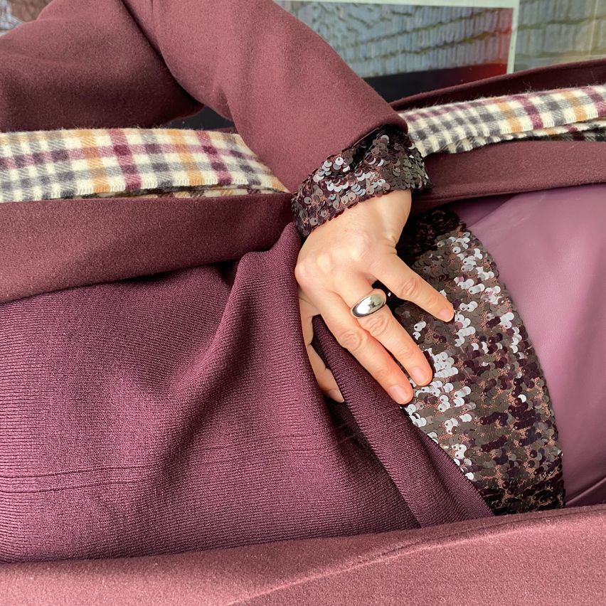 Eine Person trägt einen lilafarbenen Mantel mit einem Pailletten-Armband und einem Ring. Der Mantel hat ein kariertes Muster an den Ärmeln.