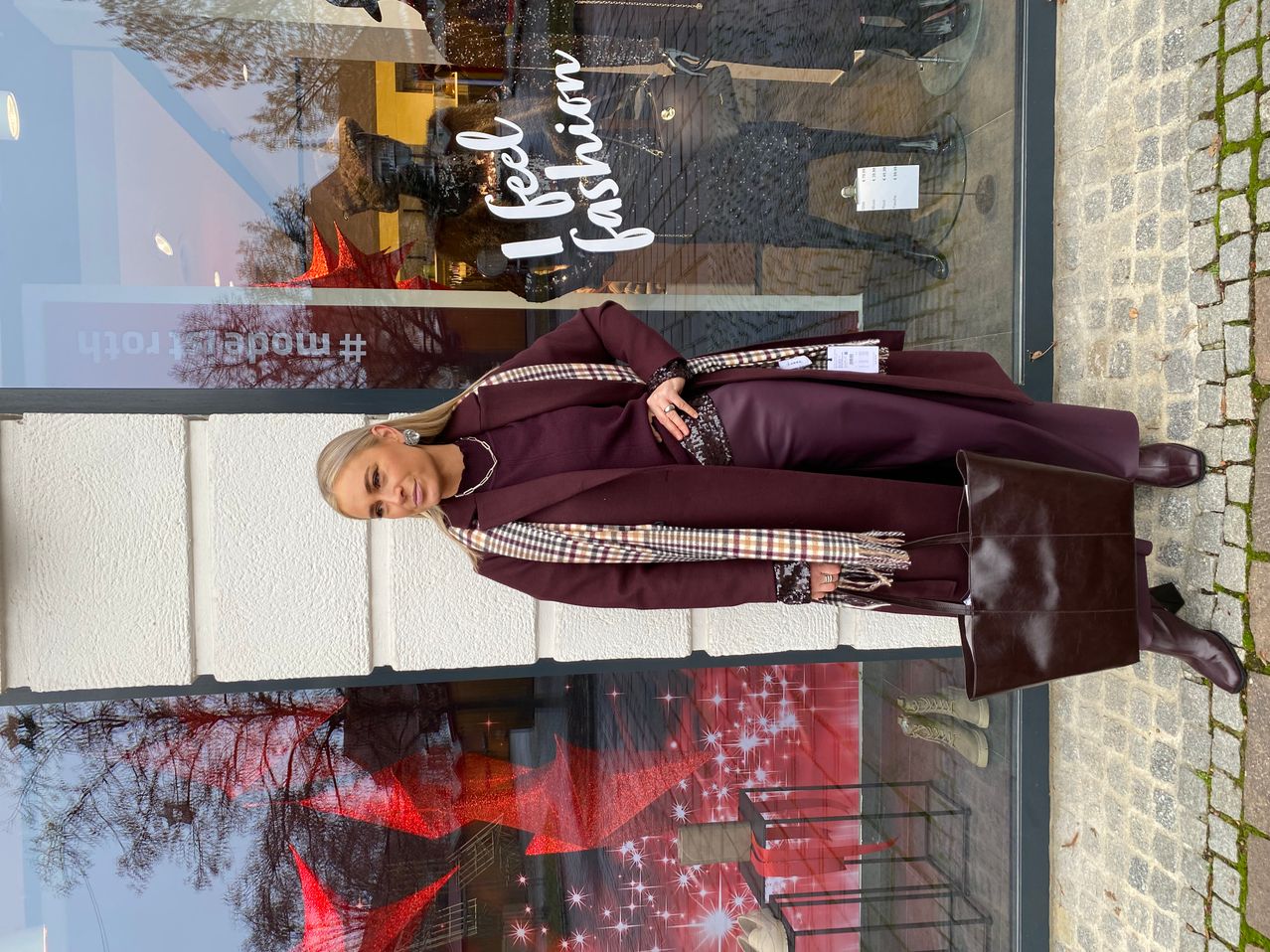 Eine Frau steht vor einem Modegeschäft mit einer Fensterdekoration, auf der 'I feel fashion' steht. Sie trägt einen Weinroten Mantel und hält eine braune Lederhandtasche. Im Hintergrund sind Reflexionen von Bäumen und roten Sternen zu sehen.