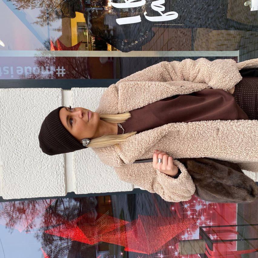 A woman with blonde hair stands outside a store with a sign that reads 'Welcome'. She wears a brown shirt, a fur coat, and a beanie. She holds a fur bag and looks at the camera.