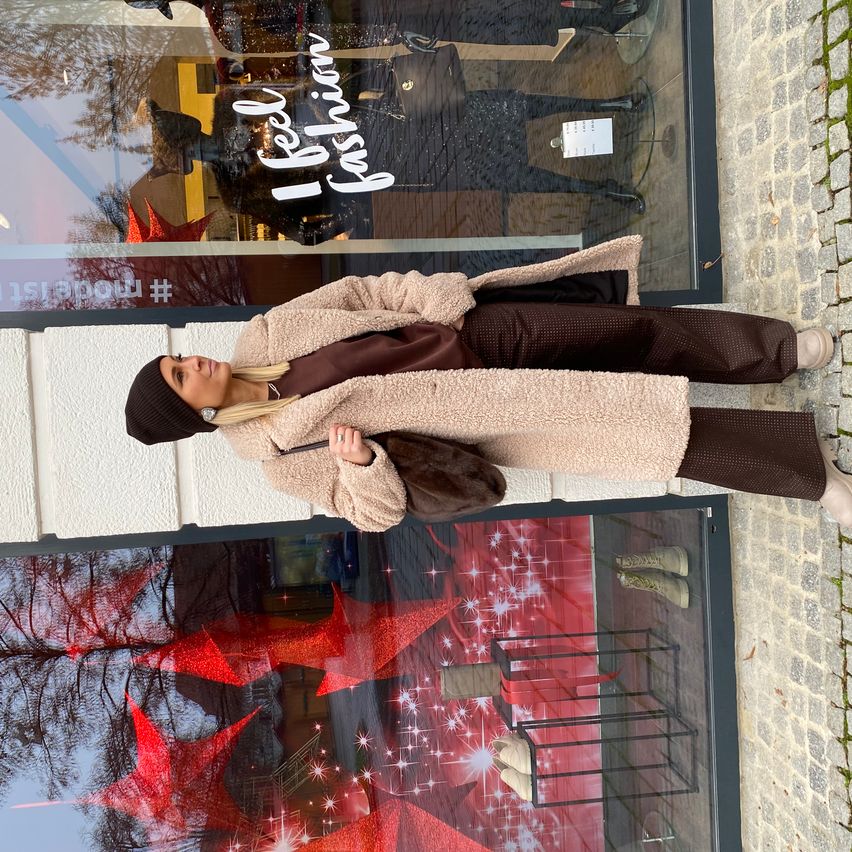 A woman wearing a brown coat and hat stands outside a shop with festive decorations, her reflection visible in the window.