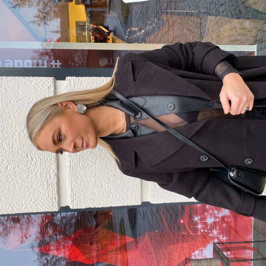 Eine Frau mit blonden Haaren posiert für ein Foto, trägt einen schwarzen Mantel und eine schwarze Tasche. Sie schaut mit einem Lächeln in die Kamera. Der Hintergrund besteht aus einer weißen Wand und einem Glasfenster mit Reflexionen.