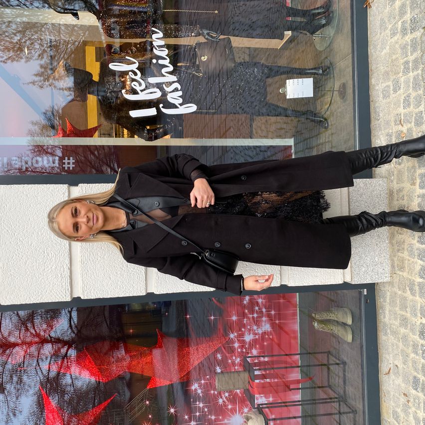 Eine Frau in einem schwarzen Mantel und Stiefeln steht vor einem Schaufenster mit roten Dekorationen.