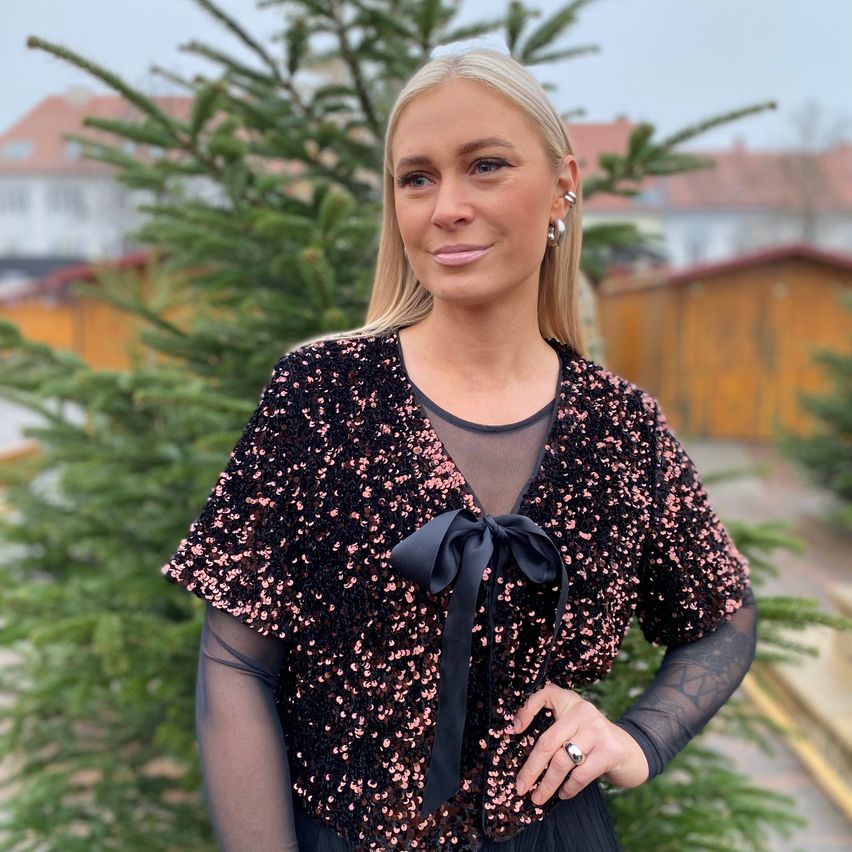 Eine Frau mit blonden Haaren steht vor einem Weihnachtsbaum und trägt ein schwarzes Pailletten-Top mit einer Schleife. Sie lächelt für die Kamera.