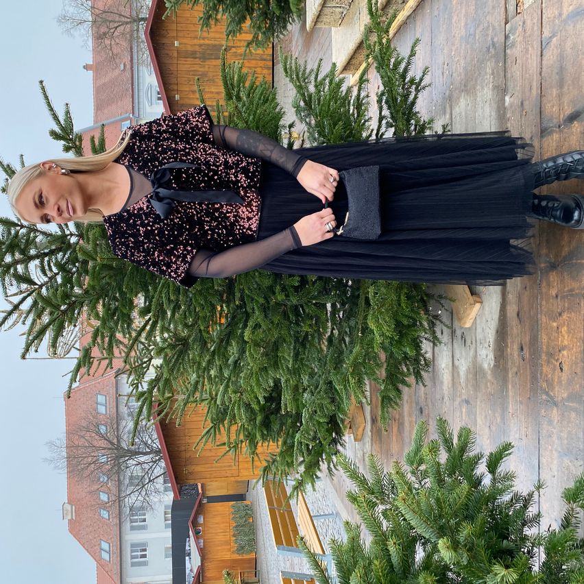 Eine Frau in einem schwarzen Pailletten-Oberteil und einer schwarzen Tüll-Skirt steht neben einer Holzwand mit Weihnachtsbäumen. Sie hält eine schwarze Clutch und ist von einem Haus mit Ziegelwänden umgeben.