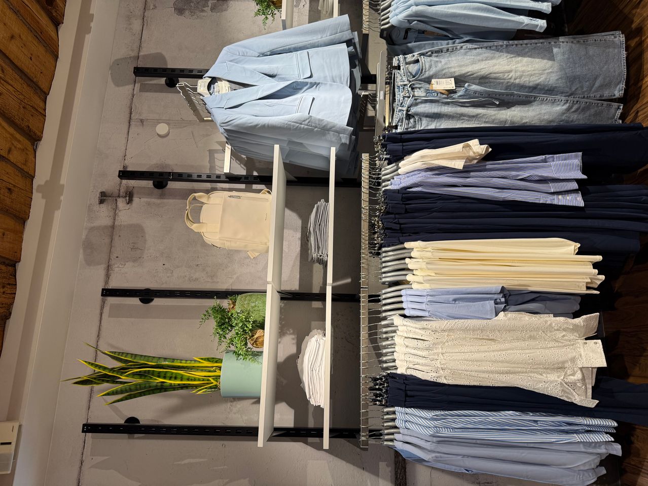 A clothing store displays various apparel on racks. A blue coat is on a hanger. Below, a beige backpack rests on a shelf. The right side shows rows of folded jeans and shirts. A potted plant with green leaves is positioned at the bottom left.