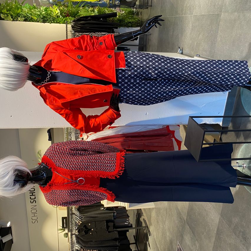 Two mannequins with white wigs display a red jacket with buttons and blue patterned pants on the left, and a red knitted jacket with fringes and blue pants on the right.
