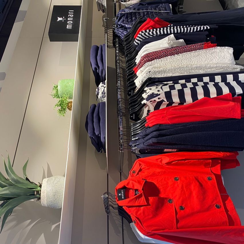 A clothing store displays various jackets and pants, predominantly in blue and red. The clothing is neatly organized on black hangers, with a black sign reading 'monari' at the top left.