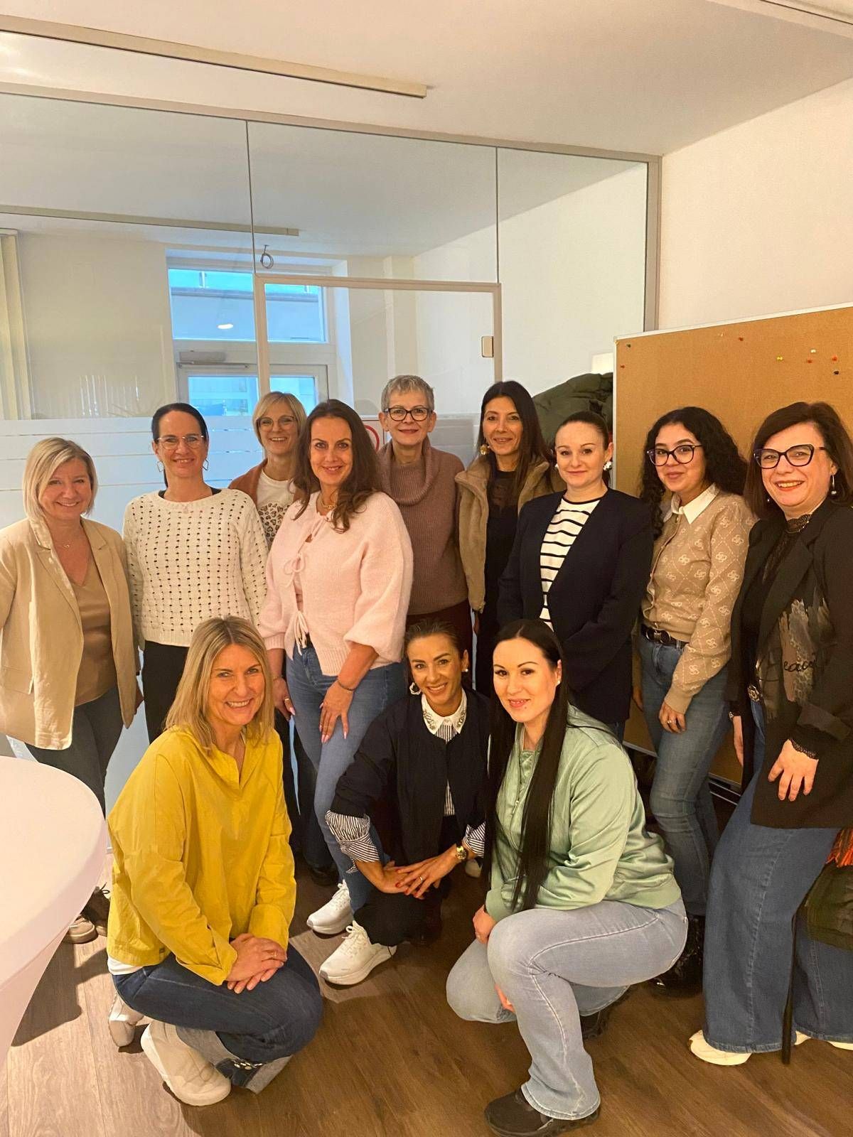 A group of women dressed in casual clothing, smiling and posing for a photo in an office. A whiteboard and table are in the background.