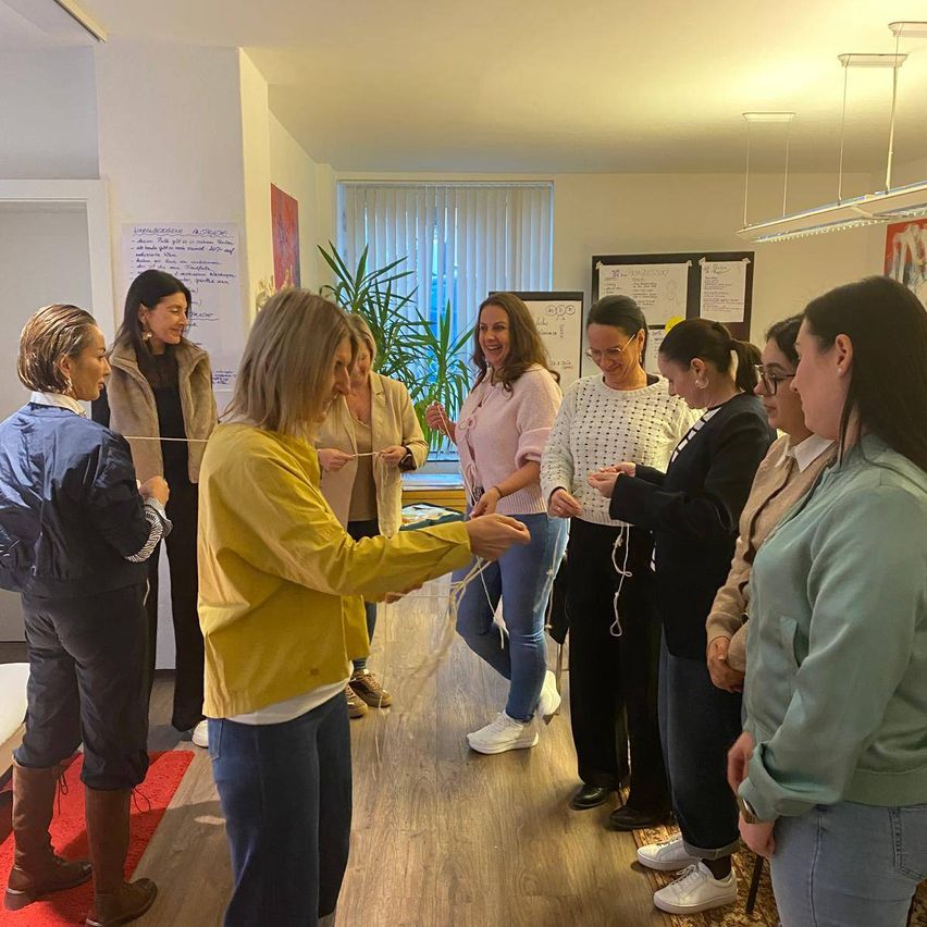 A group of women are gathered in a room, with some holding strings. A woman in a yellow jacket is holding a string in front of them. Behind them is a whiteboard, a potted plant, and a window with blinds.