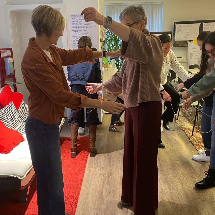 A group of people are engaged in an activity indoors. A woman is stretching her arms while another holds a string. Others are seated in chairs, participating. The room is furnished with a couch, pillows, and a carpet. Papers are attached to the wall.