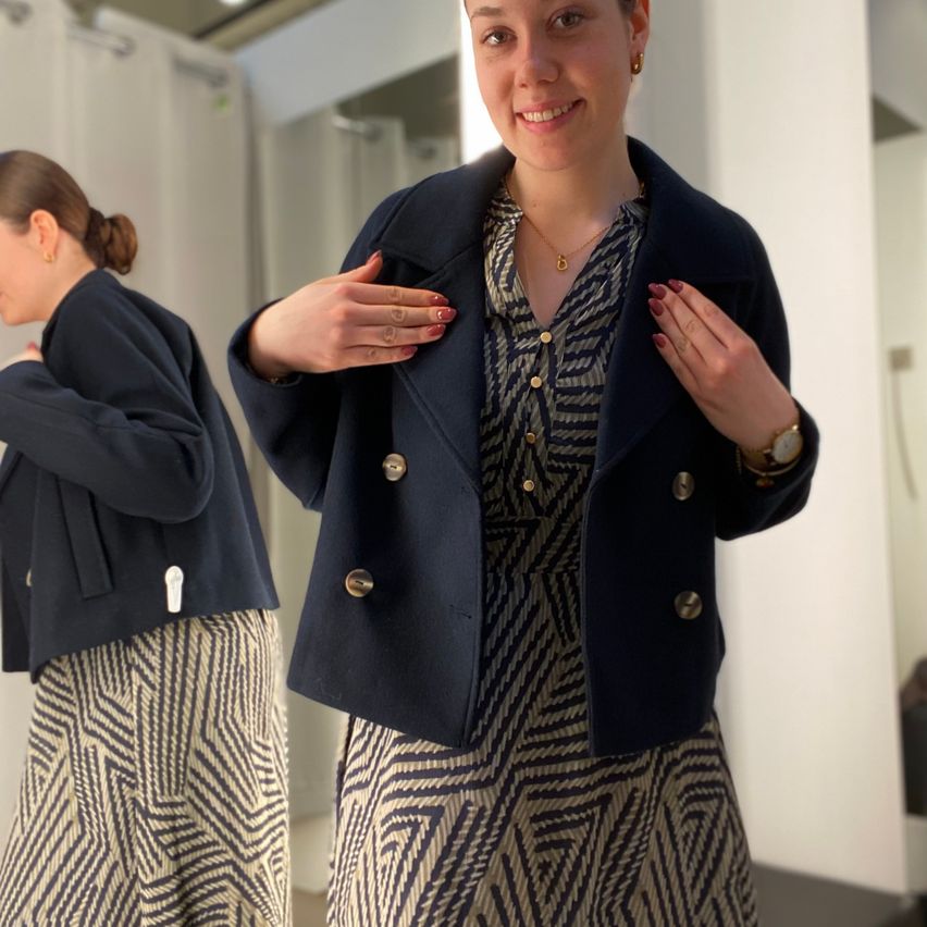 Eine Frau lächelt und posiert für ein Foto, trägt einen marineblauen Mantel mit goldenen Knöpfen und ein gemustertes Kleid. Eine andere Frau spiegelt sich im Hintergrund im Spiegel wider.
