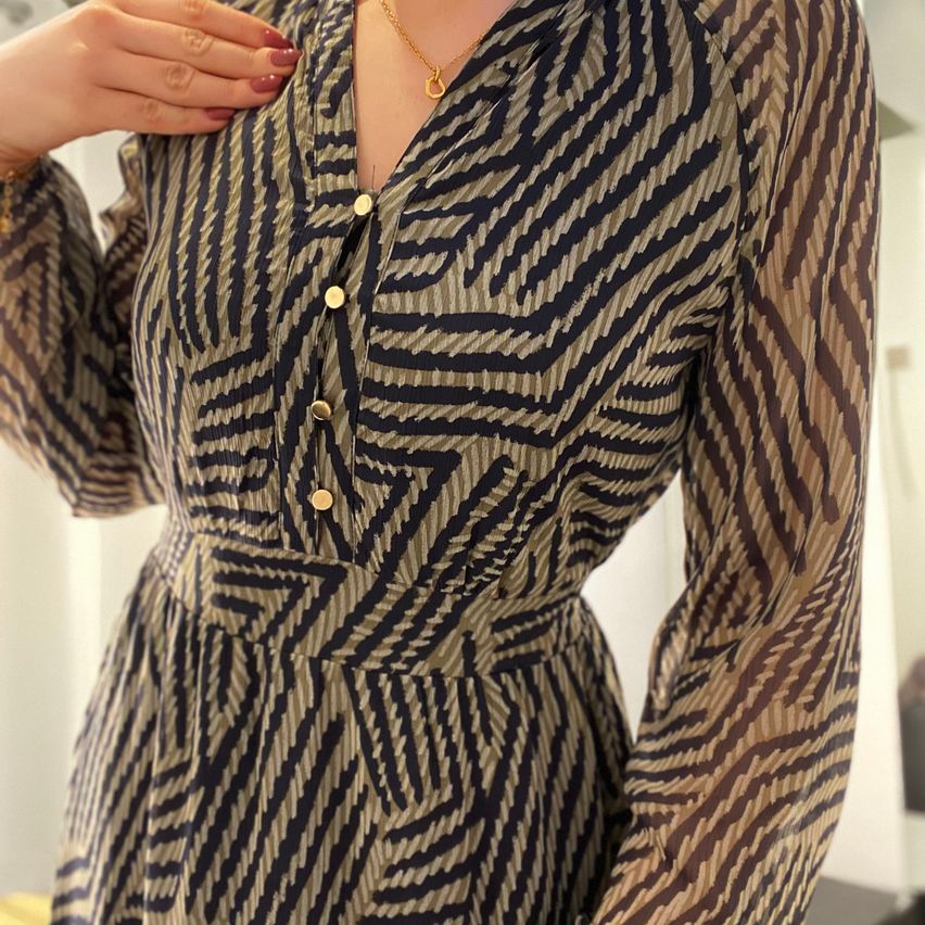 Eine Person trägt ein braun-schwarzes Kleid mit Zickzack-Muster und goldenen Knöpfen. Das Kleid hat lange Ärmel und die Person justiert es.