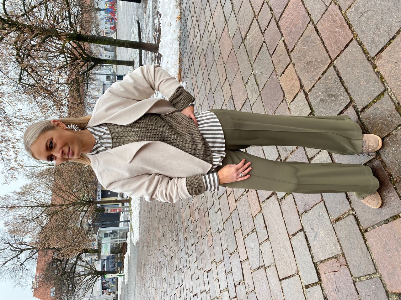 Eine Frau steht lächelnd mit den Händen auf den Hüften auf einem Kopfsteinpflasterweg und trägt einen beigefarbenen Mantel, ein gestreiftes Hemd und olivgrüne Hose.