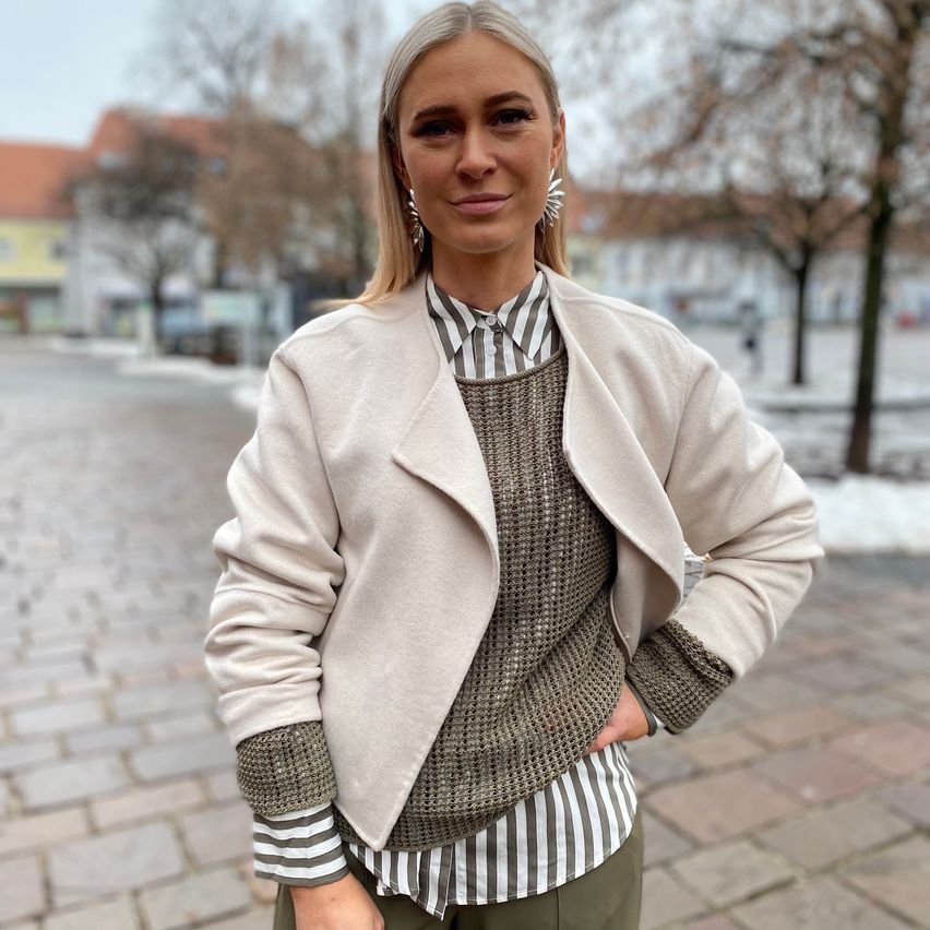 Eine Frau in einer beigefarbenen Jacke, einem gestreiften Hemd und grünen Hosen steht auf einem gepflasterten Weg mit Gebäuden und Bäumen im Hintergrund.