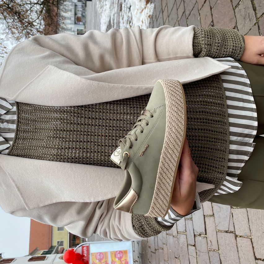 Eine Person steht draußen auf einem gepflasterten Bürgersteig und hält ein Paar olivgrüne Turnschuhe. Die Person ist in einem beigen Cardigan, einem gestreiften Hemd und grünen Hosen gekleidet. Hinter ihr befinden sich Gebäude und ein rotes Objekt im Hintergrund.