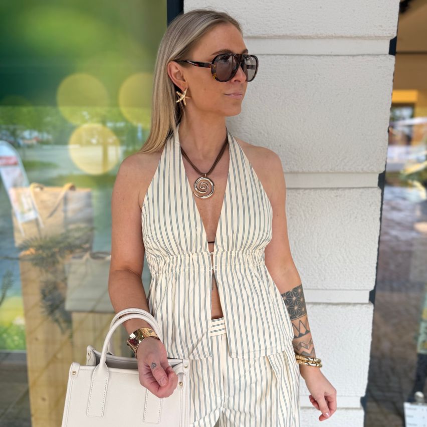 Eine blonde Frau in einem weißen und grau gestreiften Jumpsuit posiert draußen. Sie trägt eine Sonnenbrille und eine weiße Handtasche. Hinter ihr befindet sich eine weiße Wand und ein verschwommenes Ladenlokal.