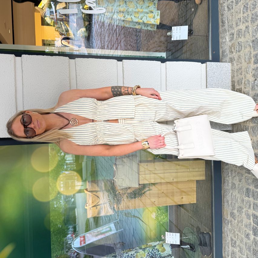 Eine Frau in einem gestreiften Jumpsuit mit Sonnenbrille posiert für ein Foto vor einem Boutique mit Glaswänden.
