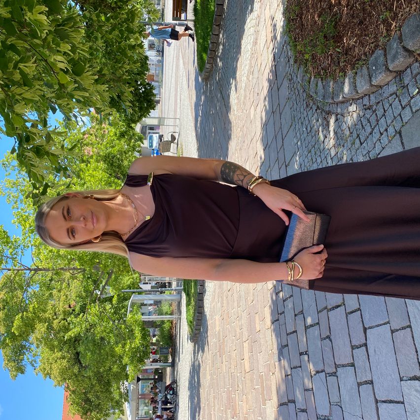 Eine Frau mit blondem Haar und braunem Kleid steht auf einem gepflasterten Gehweg und hält eine Handtasche. Im Hintergrund sind Bäume und Gebäude zu sehen.
