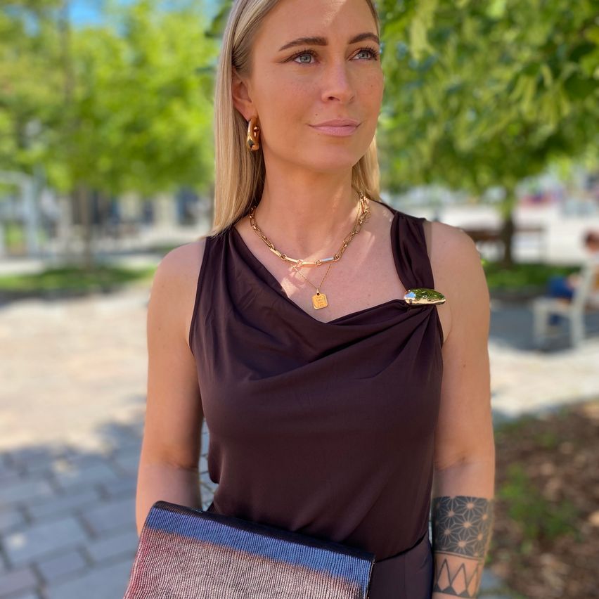 Eine Frau mit blonden Haaren, die ein braunes ärmelloses Top, eine goldene Halskette und Ohrringe trägt, steht draußen. Sie hält eine blaue und rosa Handtasche.