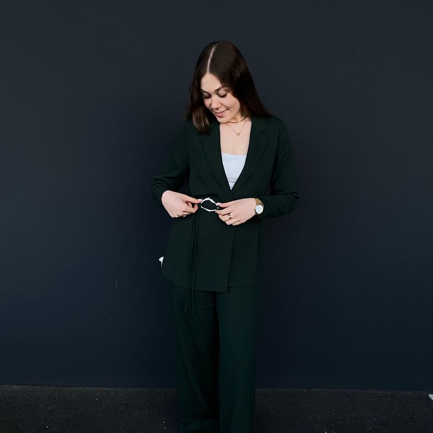 A woman in a dark green suit stands in front of a dark background. She is smiling and looking to her left, holding a pair of white eyeglasses.