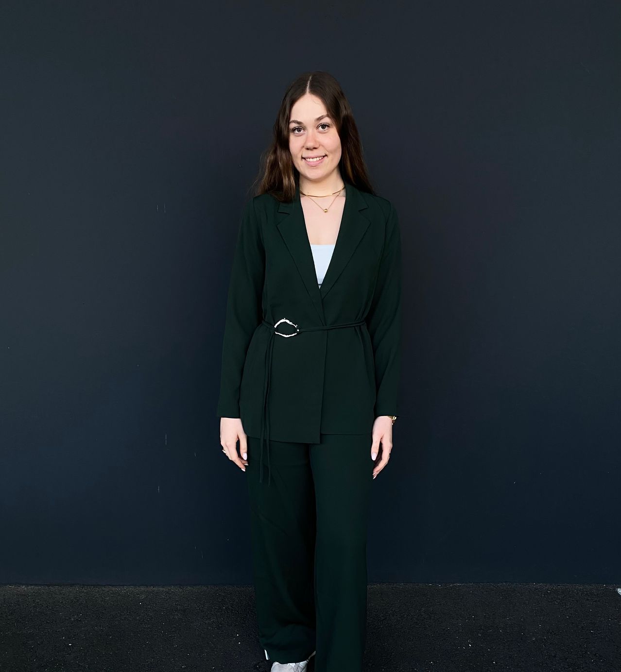 A smiling woman in a dark green suit stands against a black wall, her hands resting on her hips. She wears a light blue top, a necklace, and a silver belt.