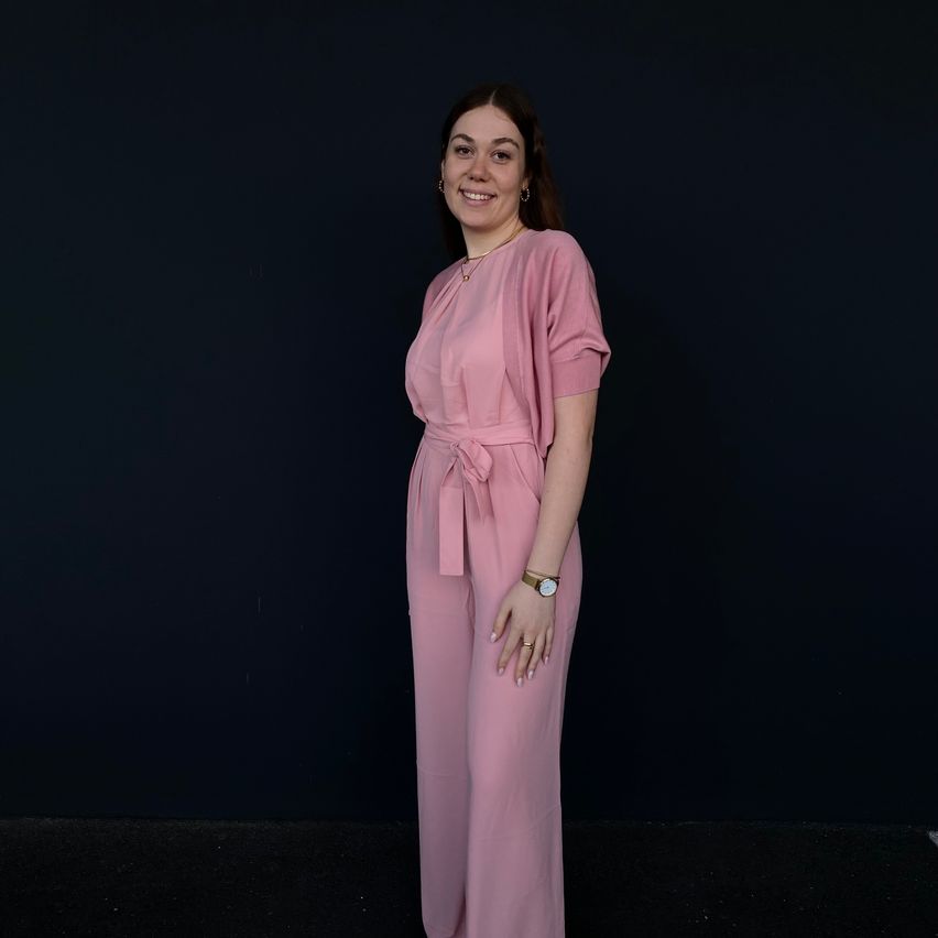 A woman in a pink jumpsuit with a belt around her waist is standing against a dark background and smiling.