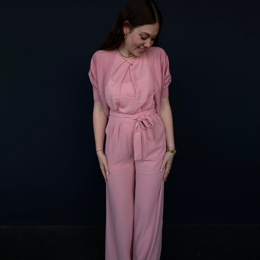 A woman stands smiling, wearing a pink jumpsuit with a belt around her waist and a necklace with a pendant. She has her hands on her hips and is looking slightly upwards.