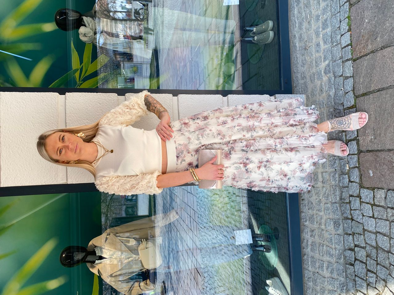 Eine Frau posiert in einem Blumenrock und weißer Bluse. Sie hat Tattoos an den Armen und hält eine kleine Handtasche. Sie steht auf einem gefliesten Boden vor einem Schaufenster.
