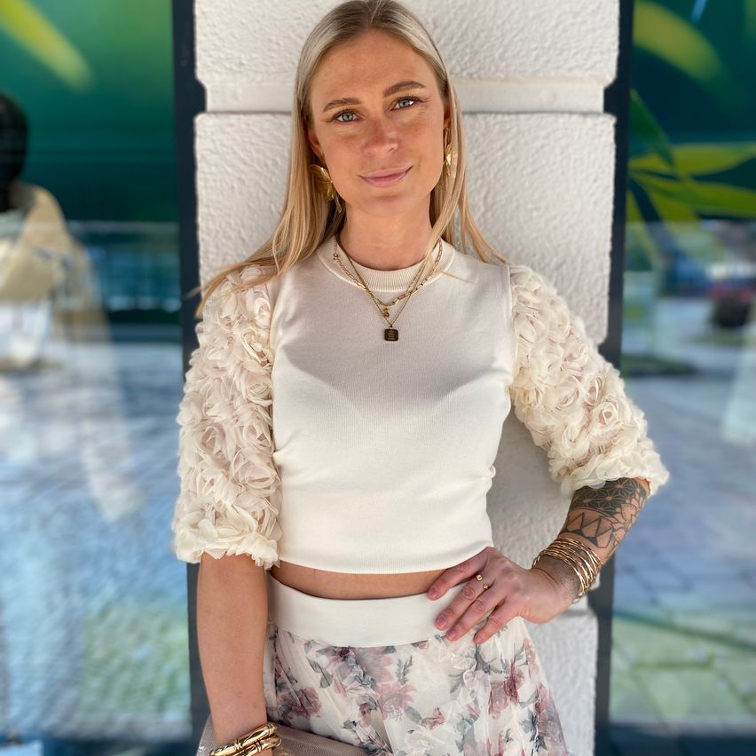Eine Frau mit blonden Haaren und Tätowierungen am Arm steht gegen eine weiße Wand und trägt eine weiße Bluse mit rüschenbesetzten Ärmeln, einen Blumenrock und eine Halskette.