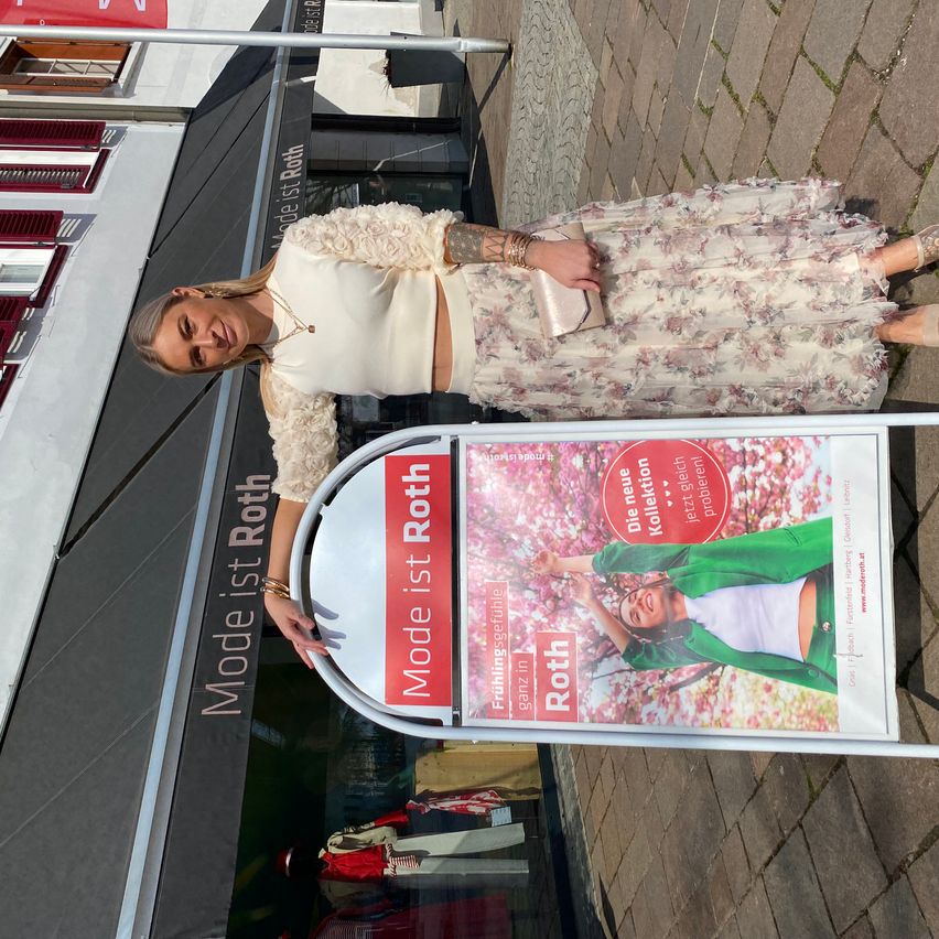 Eine Frau posiert für ein Foto vor einem Ladenschild, das 'Model ist Roth' steht. Der Hintergrund zeigt einen gepflasterten Gehweg und ein Gebäude mit einem Schild über dem Eingang.