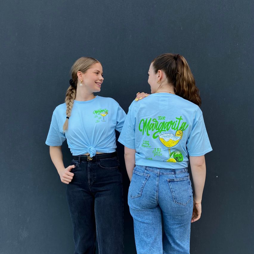 Zwei Frauen stehen Seite an Seite vor einer dunklen Wand, beide tragen hellblaue T-Shirts mit einem farbenfrohen Getränkegrafik. Eine hat eine geknotete Frisur und die andere eine Pferdeschwanz.