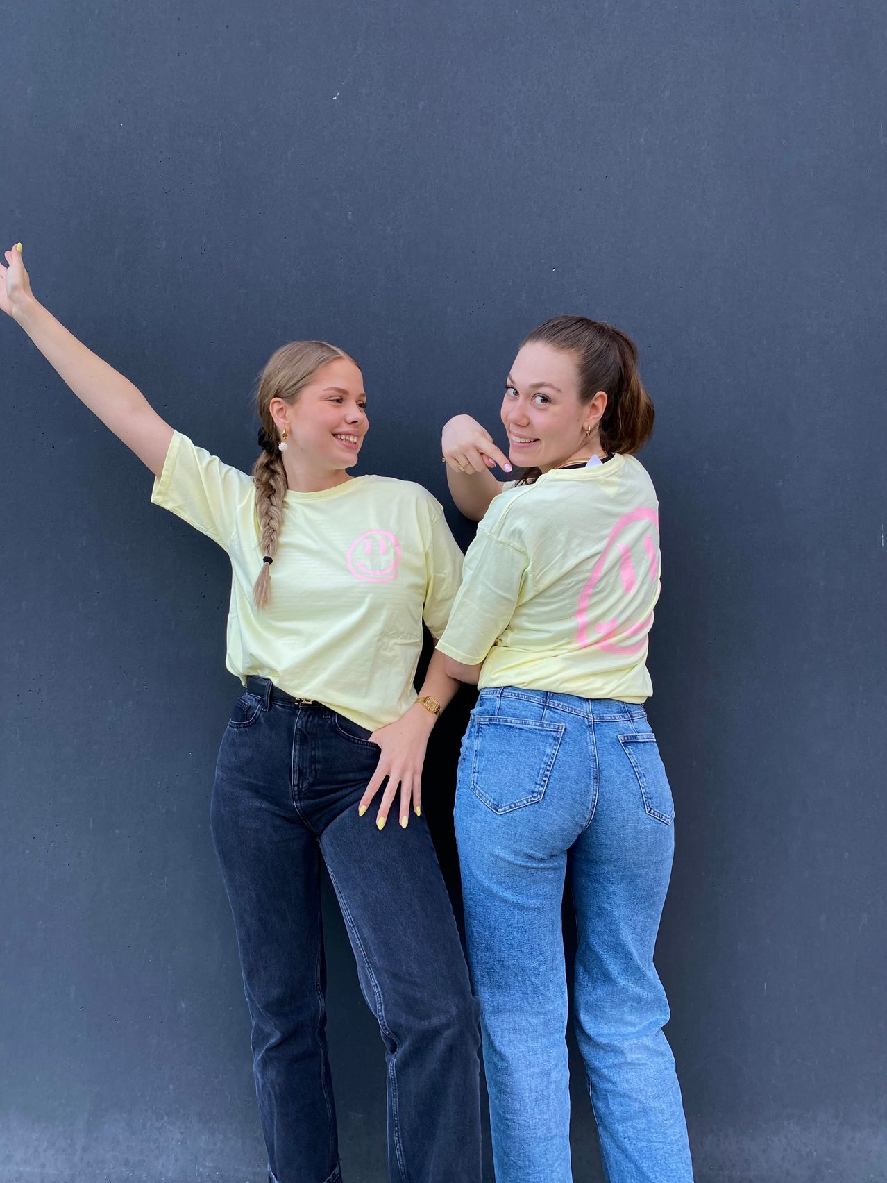 Zwei Frauen stehen an einer Wand und lächeln für ein Foto. Eine trägt ein gelbes T-Shirt mit einem rosa Grafik, die andere hat blaue Jeans und ein gelbes T-Shirt an.